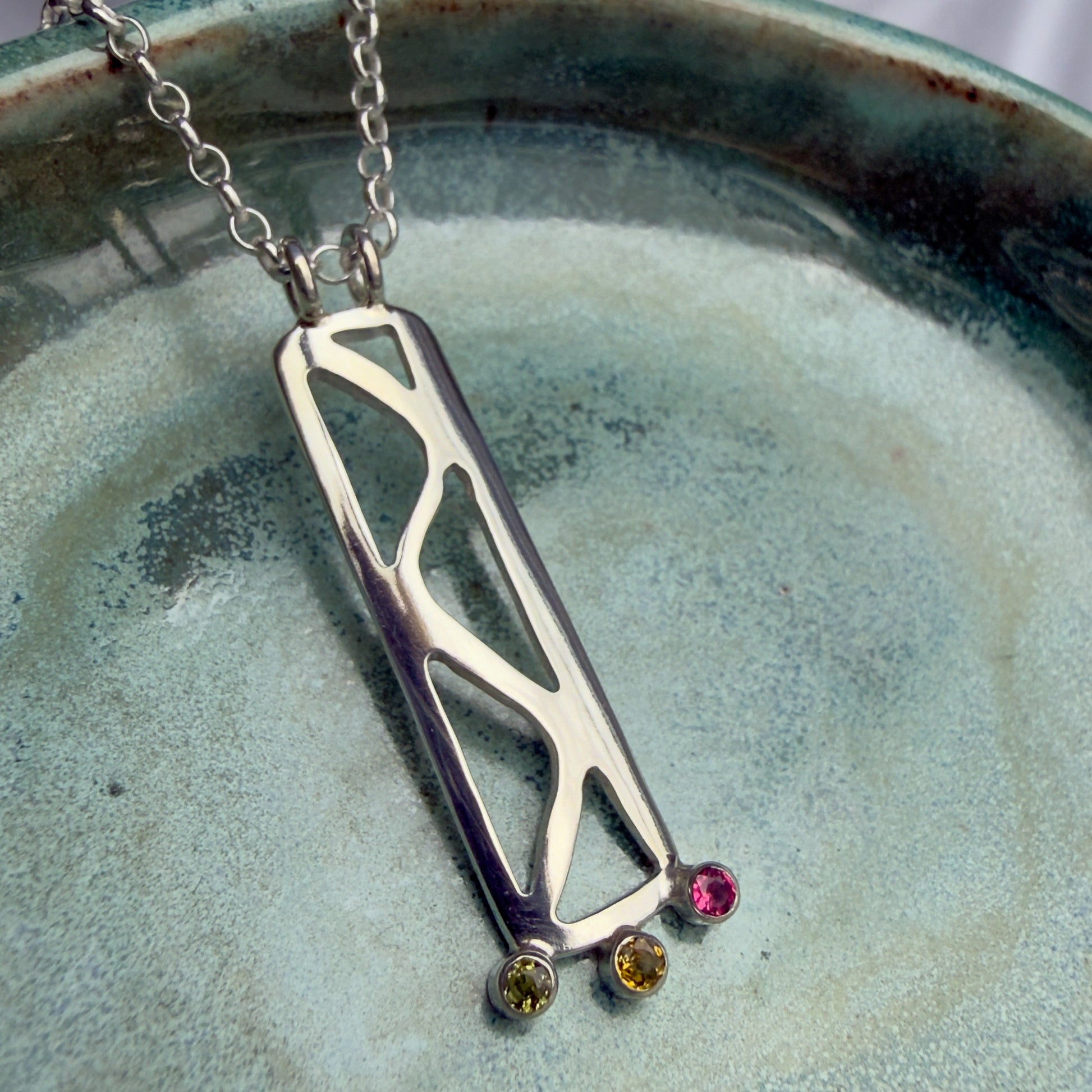 Silver necklace with a geometric pendant and three colorful stones on a ceramic plate.