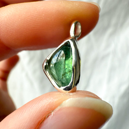 Green tourmaline gemstone pendant held between fingers against a blurred background