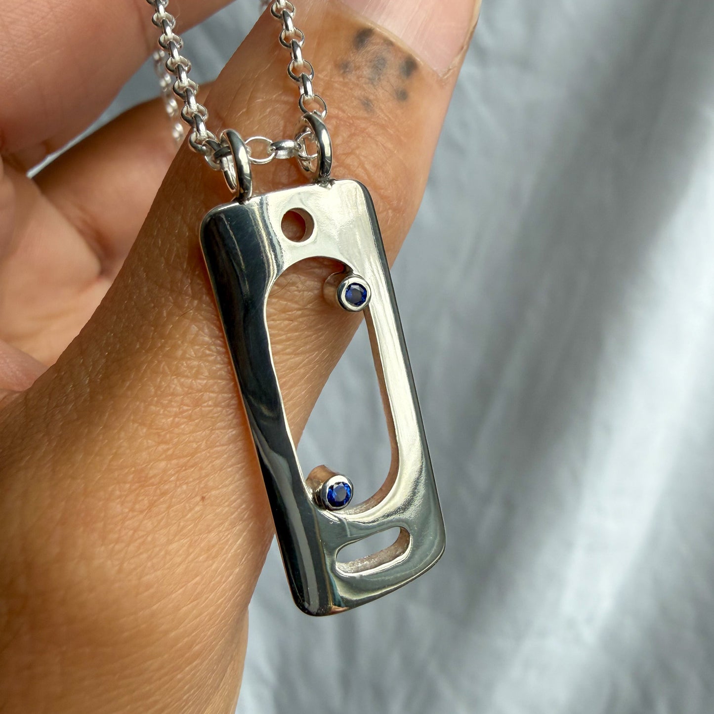 Silver pendant with a blue sapphire stone held in a hand against a neutral background