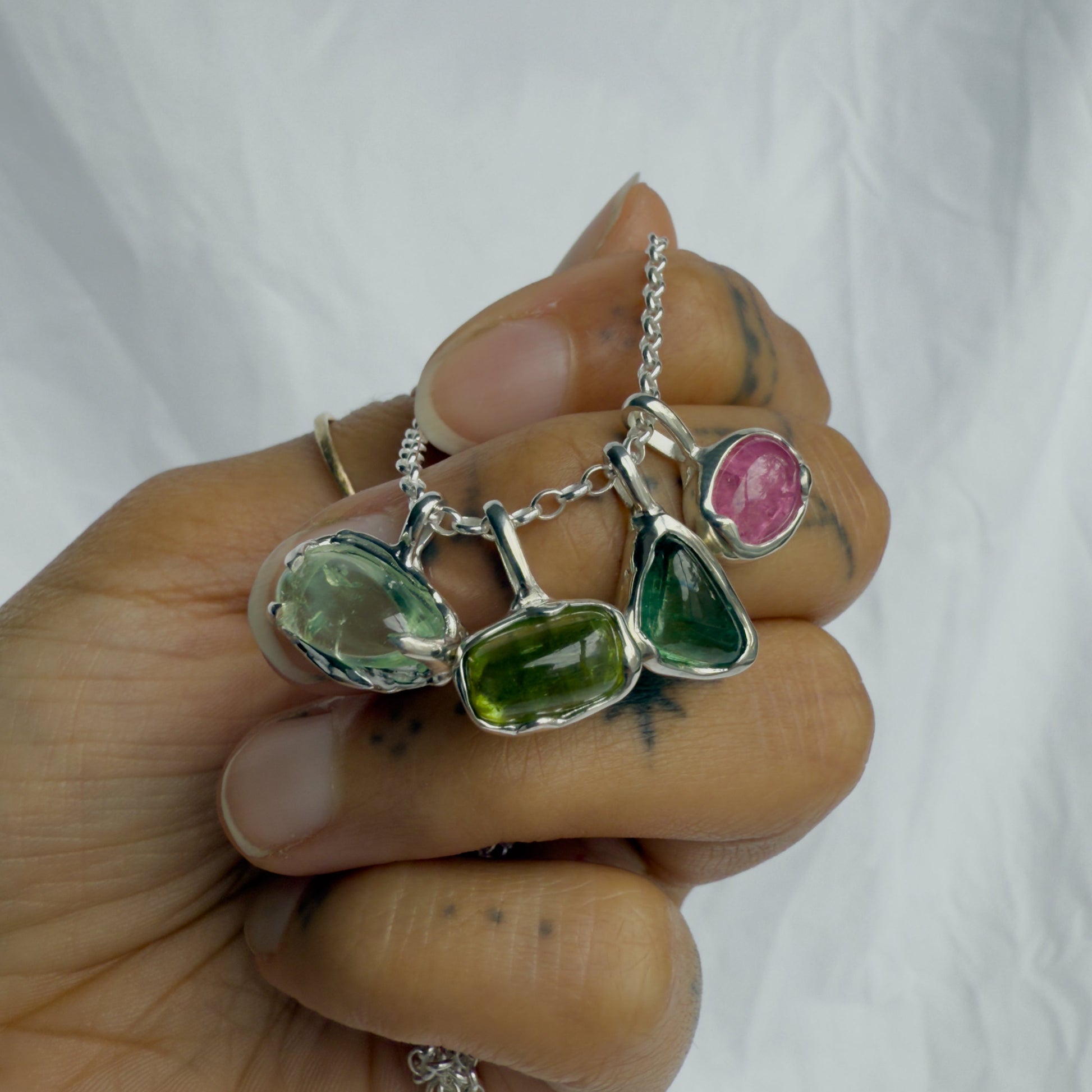 Hand holding a silver necklace with green and pink tourmaline gemstones against a white background