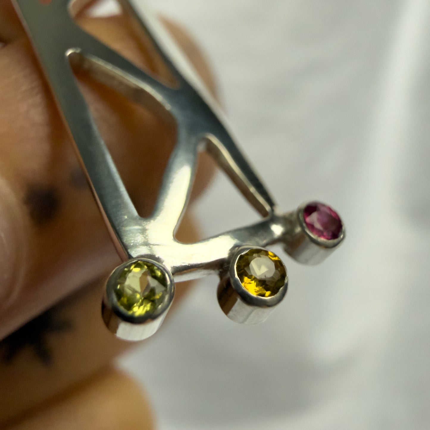 Silver pendant with three colored gemstones held by a hand against a white background